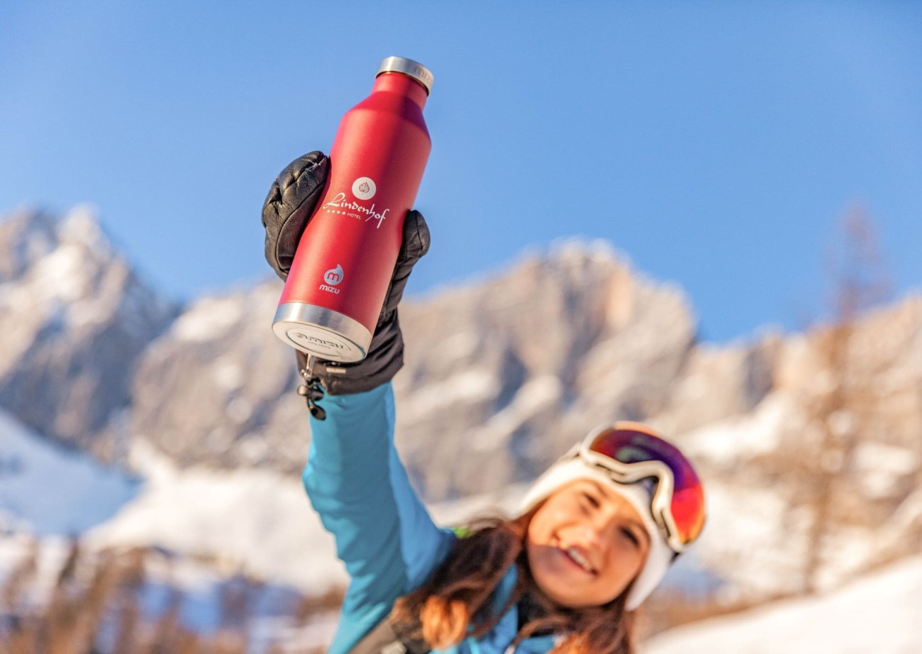Smiling woman holds up the Lindenhof water bottle surrounded by snow-covered mountains and a clear blue sky